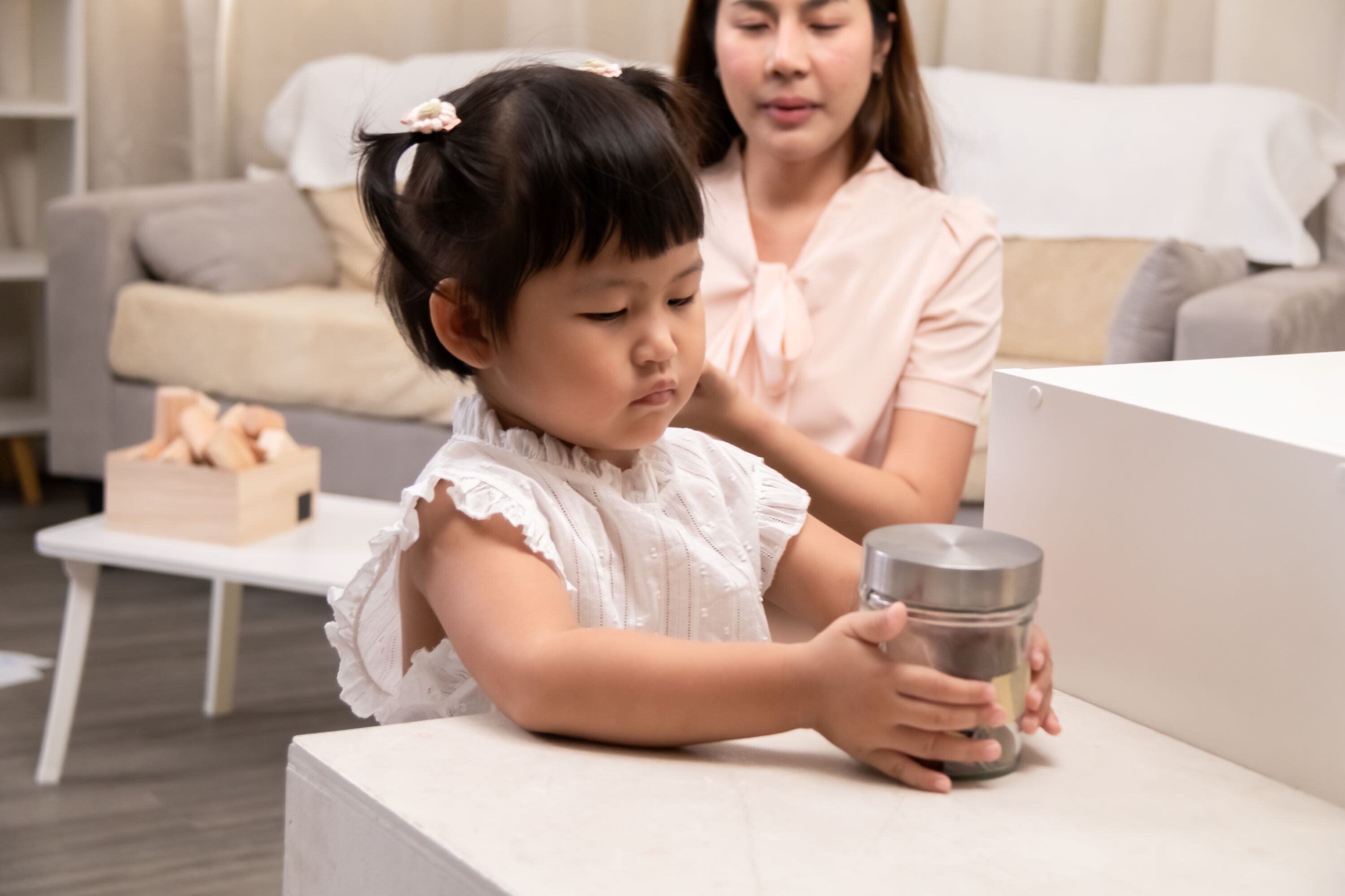 Asian toddler girl holds on to her money jar marked for tuition fees as her mom looks on.