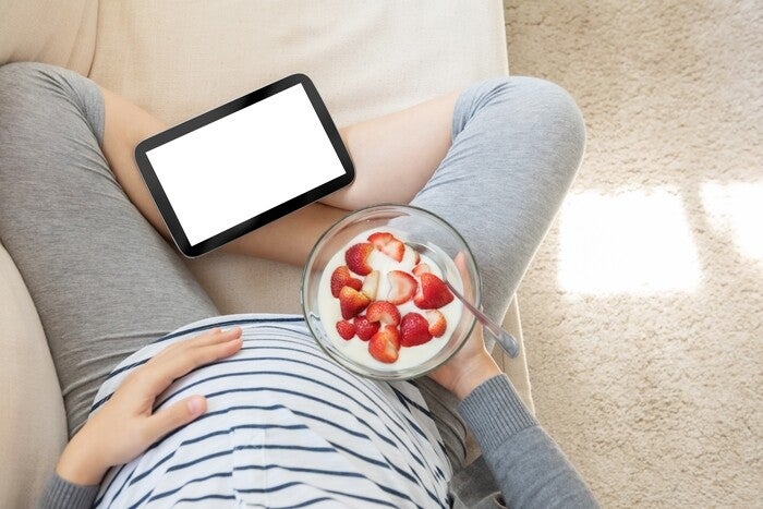 Top-view photo of pregnant woman holding a bowl of yogurt with strawberries on the sofa.