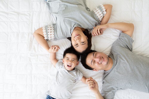 Top-view of Asian mom, dad, and toddler son lying in bed, holding hands, and smiling