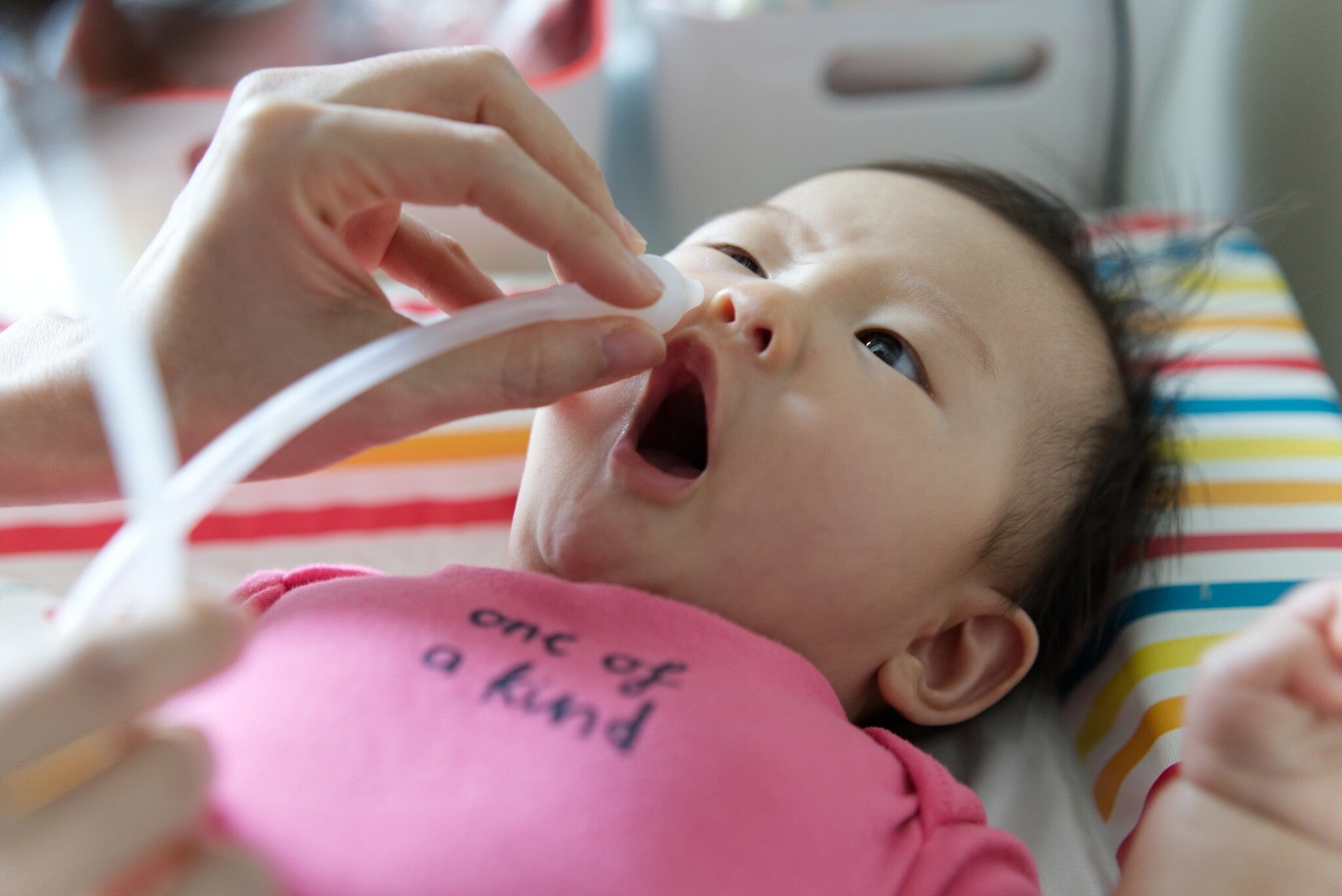 Cropped photo of woman’s hand holding a nasal aspirator tube to an Asian baby’s nose.
