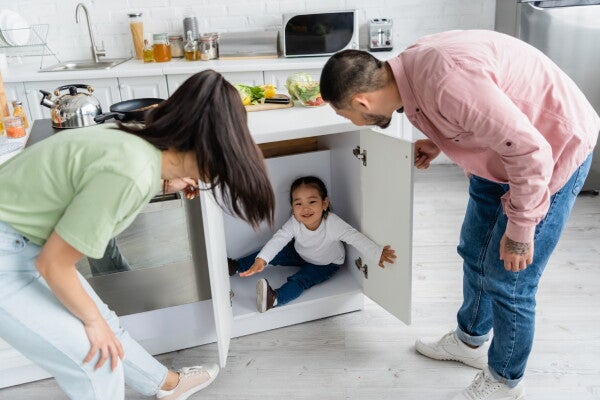 A young Asian couple finds their toddler girl hiding inside the bottom kitchen cabinet.
