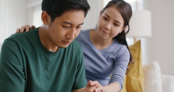 An Asian woman listens and comforts her worried-looking husband.