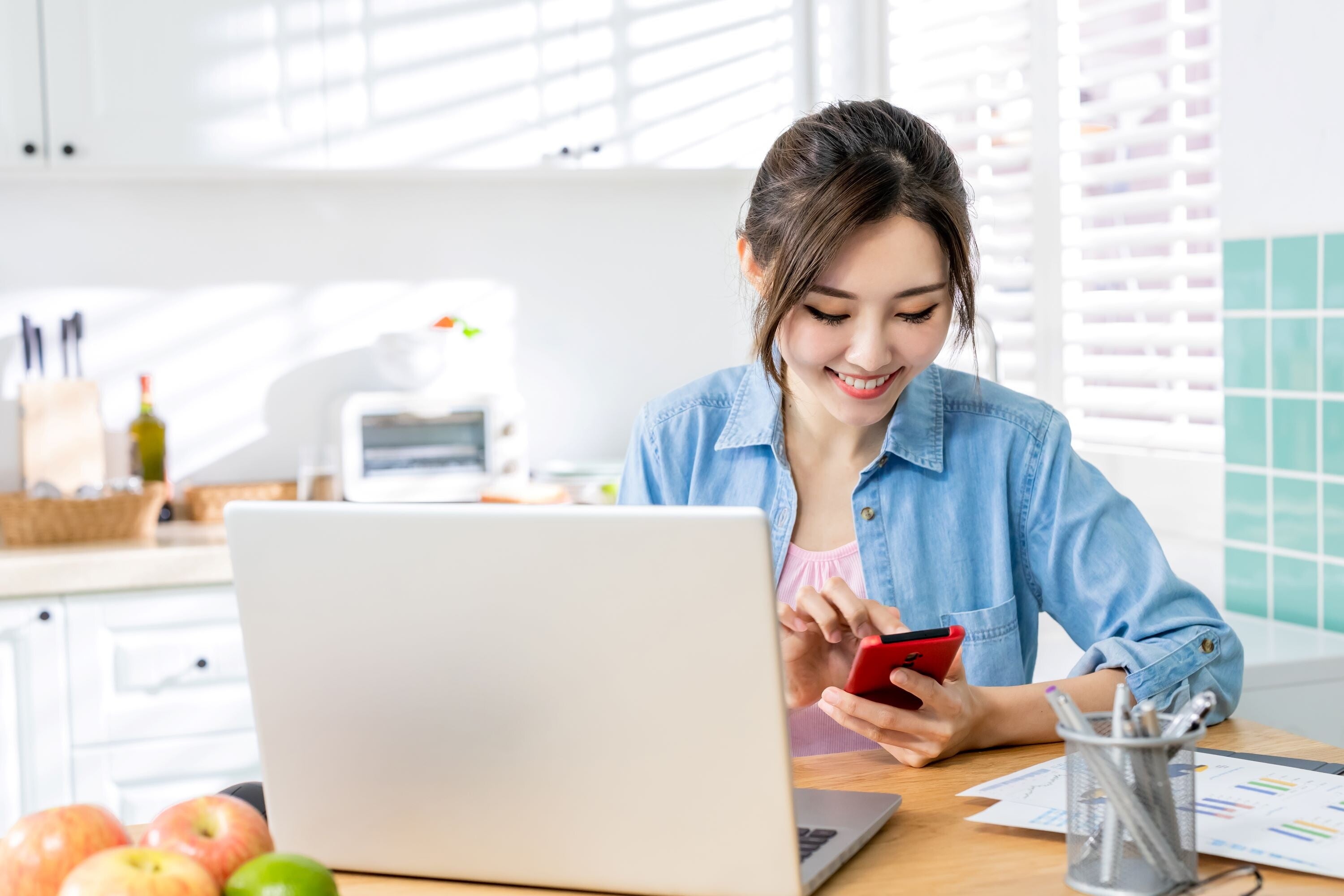 A young Asian woman sitting at a kitchen table with papers, a laptop, and a phone, looking happy.