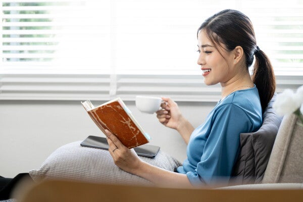 A young Asian woman relaxing, reading a book and drinking coffee in her living room.