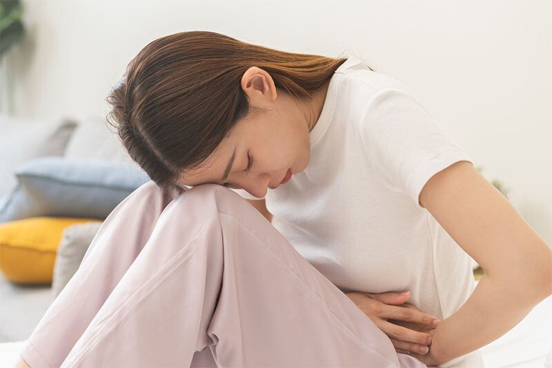 Young Asian woman sits in bed with knees up to her chest as she holds her belly in pain