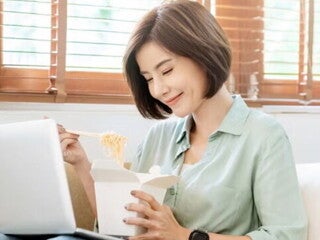 Young Filipina experiences a food craving and enjoys takeout while sitting on the sofa with her laptop on her lap.