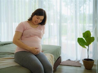 Happy pregnant Asian woman touches her pregnant belly while sitting on the bed. Happy pregnant Asian woman touches her pregnant belly while sitting on the bed.