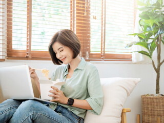 Young Filipina experiences a food craving and enjoys takeout while sitting on the sofa with her laptop on her lap. Young Filipina experiences a food craving and enjoys takeout while sitting on the sofa with her laptop on her lap.