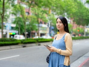 A 17-week-pregnant Asian woman holding her smartphone while standing on the street.