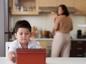 A little Asian boy watching a show on a tablet while his mother is cooking dinner in the background A little Asian boy watching a show on a tablet while his mother is cooking dinner in the background
