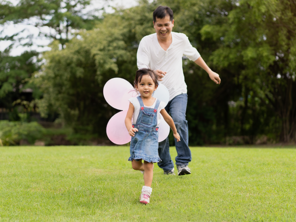 A smiling Asian preschooler girl running in the park with her dad for a budget-friendly day out. 
