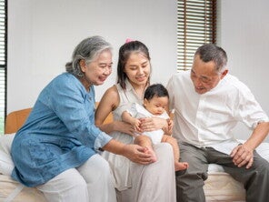 Asian grandparents look lovingly at their grandchild who is held by their mother on her lap.