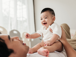 Asian dad lying on bed talks to his smiling baby who's sitting on his stomach Asian dad lying on bed talks to his smiling baby who's sitting on his stomach
