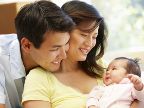 Asian father embraces his wife as she carries their infant who smiles at them, a new step in the child development stages.