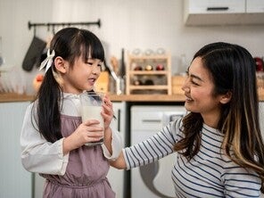 Asian preschooler girl with a glass of milk as her mom smiles at her in the kitchen, happy she’s getting protein in milk