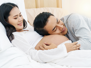 Asian woman who is 22 weeks pregnant lie in bed while her husband puts his ear to her growing belly.