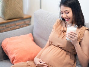 Pregnant Asian woman on the sofa, looking at and touching her belly while holding a glass of milk for pregnant women