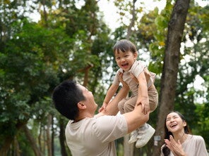 Cropped photo of a dad outdoors lifting his little boy in the air as a form of risky play that builds self-confidence in kids while mom looks on 