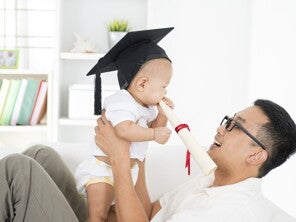 Asian dad smiles at his baby who has a graduation cap on as he bites on a diploma. 
