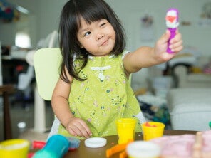 Asian preschool-age girl lifts a toy and tilts her head as she looks at it with one eye closed.
