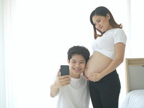 Young Asian pregnant couple taking selfie by a window for bright and beautiful baby bump pictures.