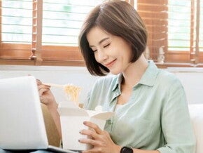 Young Filipina experiences a food craving and enjoys takeout while sitting on the sofa with her laptop on her lap. Young Filipina experiences a food craving and enjoys takeout while sitting on the sofa with her laptop on her lap.