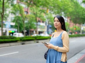 A 17-week-pregnant Asian woman holding her smartphone while standing on the street.