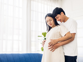 A 26 weeks pregnant woman holding her belly while her husband hugs her from behind in their home
