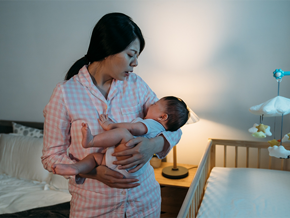 A sleepy Asian mom in pajamas carrying her newborn, who’s fussy because of colic in babies