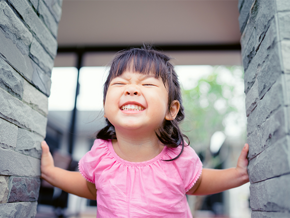 A smiling Asian toddler between two stone pillars with smiling to show her healthy milk teeth.