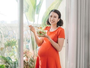 A young Asian woman standing by the window and holding a bowl of salad as part of her healthy pregnancy meal plan