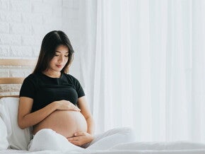 A young pregnant Asian woman holding her belly and seated in bed, alert to the signs of labor