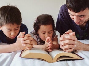 Asian dad with young son and little girl praying on the bed at home, one of their Filipino traditions.