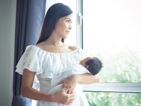 A young Asian mom holding her baby and looking out the window to see if it’s safe to open for a healthy environment.
