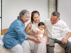 Asian grandparents look lovingly at their grandchild who is held by their mother on her lap.