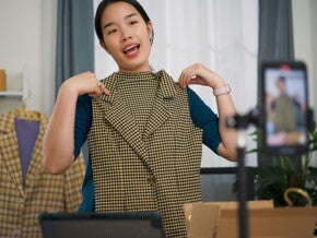 A young Asian woman sitting at a kitchen table with papers, a laptop, and a phone, looking happy. 