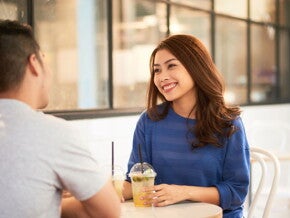 A young Asian single mom seated on a café table and smiling at her date while holding her juice.