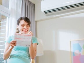 Asian woman a bright living room looking at her family’s electricity bill and smiling at savings.