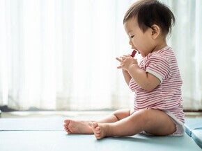 Asian baby sitting on a mat about to put a choking hazard in his mouth