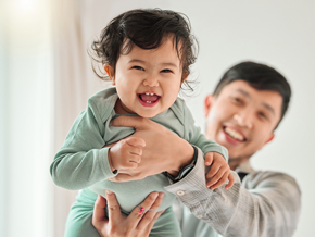 Asian dad lifts his laughing baby with both hands high in the air 
