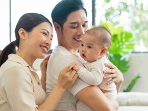 Asian dad with his wife behind him carries their healthy baby in his arms.