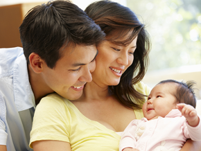 Asian father embraces his wife as she carries their infant who smiles at them, a new step in the child development stages.