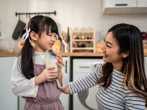Asian preschooler girl with a glass of milk as her mom smiles at her in the kitchen, happy she’s getting protein in milk