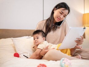 Asian working mom with her baby in bed talks on the phone while looking at documents.