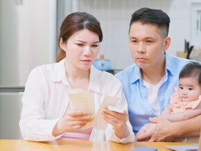 A worried Filipino couple sitting at a table with their baby and their bills, discussing finances and their source of income.