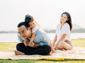 Little Asian girl hugs her dad as they spend an afternoon in the park for Father’s Day, while mom smiles and watches.