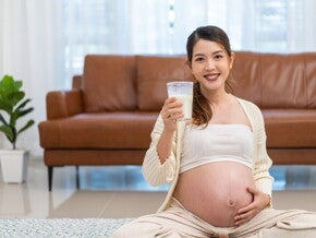 A Practical Guide to Calcium in Pregnancy Pregnant Asian woman holding a glass of milk in front of brown sofa.