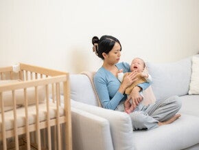 A young Asian mom carrying her little one while sitting on the sofa, wondering how to make a baby sleep.