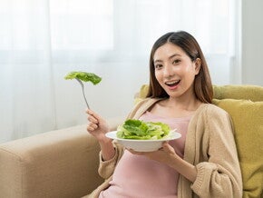 Asian pregnant woman eating greens on sofa. Asian pregnant woman eating greens on sofa.