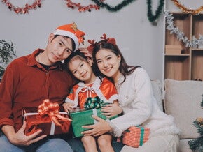 A young Asian couple with their little girl posing for a Christmas photoshoot while holding Christmas presents and wearing holiday headwear.